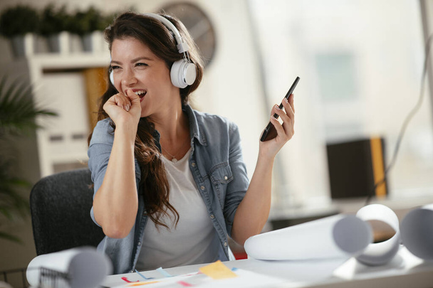 Νεαρή γυναίκα στο γραφείο με ακουστικά. Νεαρή περιστασιακή επιχειρηματίας απολαμβάνοντας το αγαπημένο της τραγούδι πάνω από ακουστικά και τραγουδώντας ενώ εργάζεται σε φορητό υπολογιστή στο γραφείο. - Φωτογραφία, εικόνα