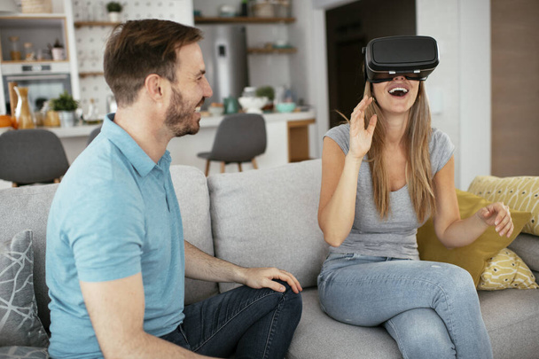 Ευτυχισμένο ζευγάρι που διασκεδάζει με γυαλιά εικονικής πραγματικότητας στο σαλόνι. Νεαρό ζευγάρι που παίζει και απολαμβάνει βιντεοπαιχνίδια με ακουστικά VR στο σπίτι. - Φωτογραφία, εικόνα
