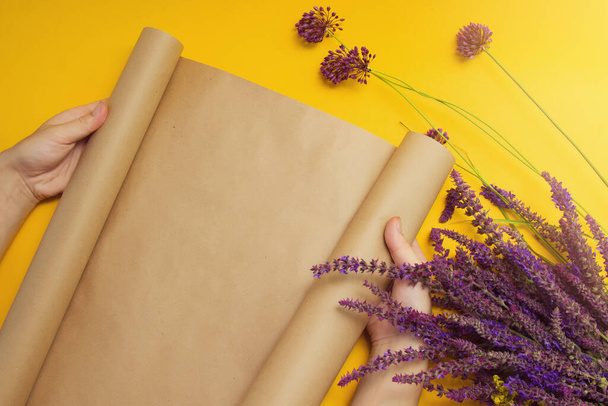 Female hands unfold craft paper next to a bouquet of lavender flowers and wild onions on a bright yellow background. Top view. Place for text. - Фото, изображение