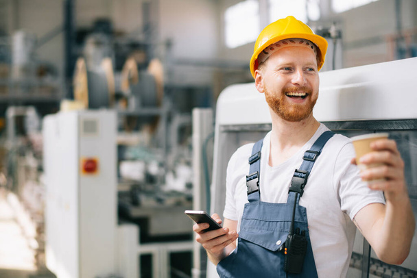 felice lavoratore in casco giallo utilizzando il telefono in fabbrica  - Foto, immagini