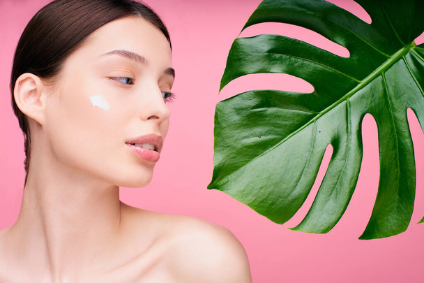 Close-up, portrait of a tender young caucasian girl with a nourishing face cream on her cheek, holding a green monstera leaf near her face, nude. Natural beauty and organic cosmetics concept. - Photo, Image