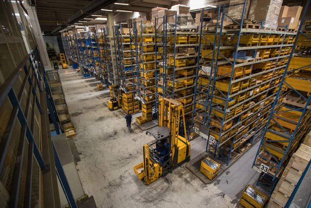 Interiér distribučního skladu shora. Pohled na modré regály a žluté krabice. Logistika a skladování v plné akci s vysokozdvižným vozíkem a spoustou zboží kolem - Fotografie, Obrázek