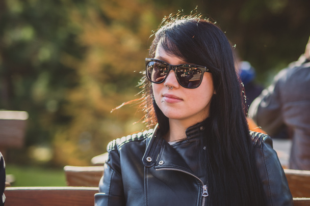 Cute sexy young woman with long black hair, sunglasses, septum piercing and a leather jacket sitting on a bench in a park and enoying the sun. Fetish style woman in leather outdoor - Photo, Image