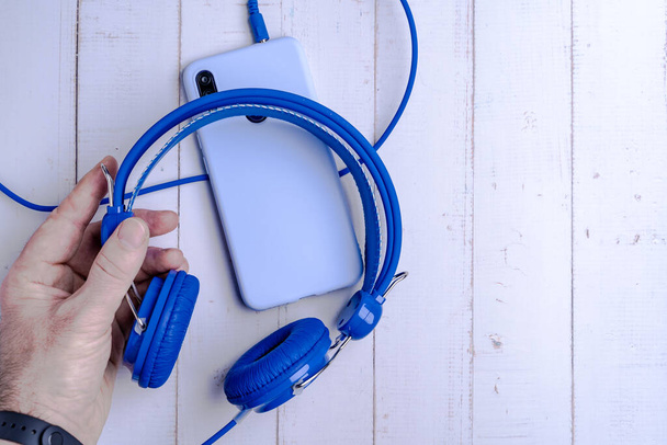 A top view of a hand holding blue headphones connected to a phone on a wooden surface for a copy space - Foto, Imagem