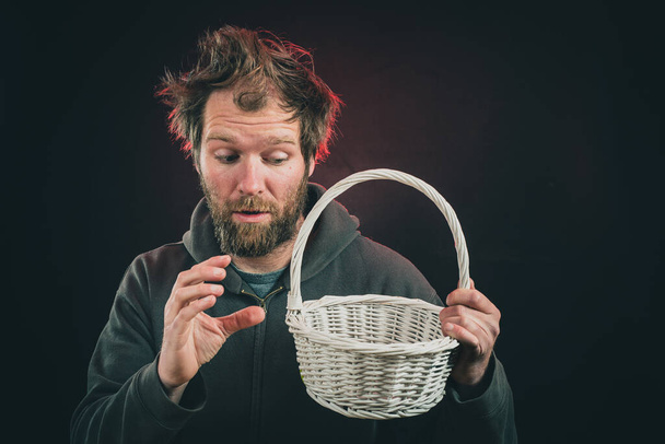Young man pretending to be curiosly lifting something from a white wowen basket on a black background and red backlight. Concept of taking something from a container. - Photo, Image