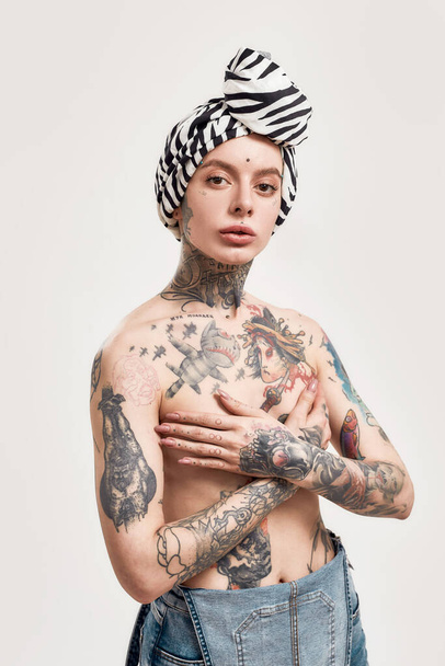 Tatuaż i kolczyk. Biała, przekłuta i wytatuowana kobieta stojąca topless z turbanem zakrywającym piersi z rękami skrzyżowanymi na klatce piersiowej w dżinsach patrząca w kamerę - Zdjęcie, obraz