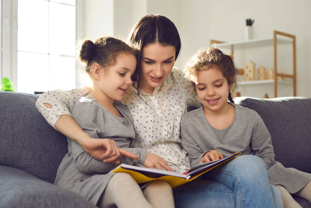 Happy young mother reading a book sitting at home on the couch with her twin daughters. - Foto, imagen