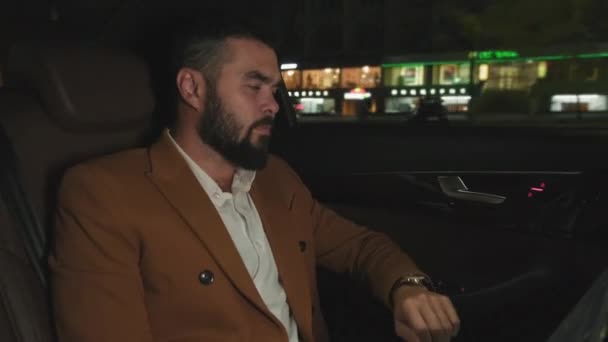 Handheld-Medium-Aufnahme eines gut aussehenden Geschäftsmannes in Kameljacke, der nachts auf dem Rücksitz eines teuren Autos sitzt und auf seine Armbanduhr schaut - Filmmaterial, Video
