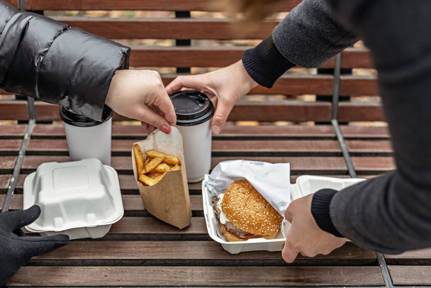 Руки женщины разложили гамбургеры с чашками кофе, картошку фри на скамейке в парке. Фастфуд. Концепция еды на вынос. - Фото, изображение