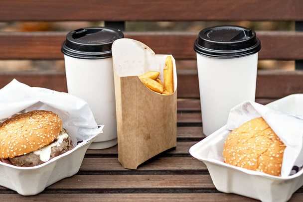 Фастфуд. Бумажные чашки с кофе, гамбургеры коробки и картофель фри на скамейке в парке.. - Фото, изображение