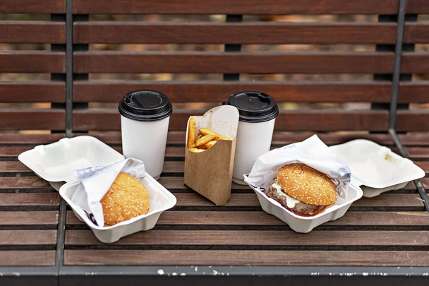 Comida rápida. Tazas de papel con café, cajas de hamburguesas y papas fritas en un banco del parque.. - Foto, Imagen