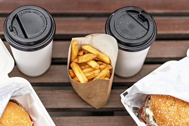 Fast food. Paper cups with coffee, hamburger boxes and fries on a park bench.Takeaway food concept. - Photo, Image