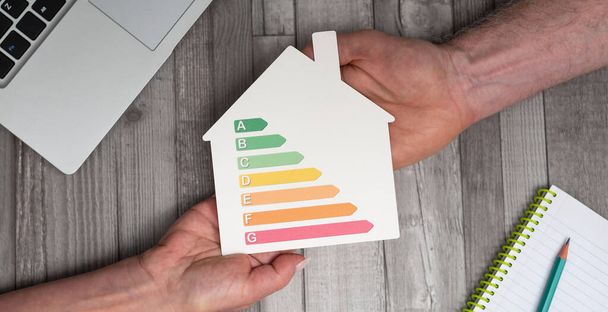 Руки, держащие в руках дом, показывающий энергоэффективность дома; Концепция оценки эффективности дома - Фото, изображение