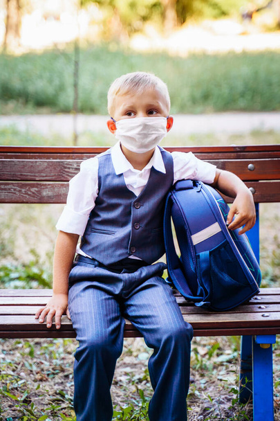Young schoolboy in a protective medical mask sits on a bench in the park in front of the school. Dressed in formal uniform, holding backpack nearby. New reality during coronavirus pandemic. Covid-19. - Photo, Image
