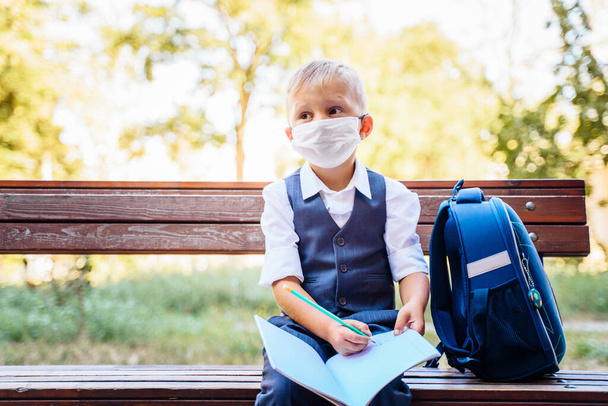 Elementary school student sits on a bench. Dressed in smart school uniform. Holding notebook on his knees and writing in it. New normal. Wears a medical protective mask for coronavirus. Covid-19. - Photo, Image