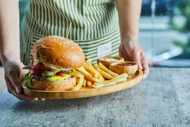 Hände halten einen Holzteller mit Burger und Bratkartoffeln mit Ketchup und Mayonnaise - Foto, Bild