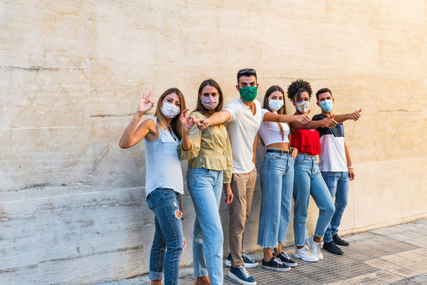 Gruppenporträt multiethnischer Jungen und Mädchen mit bunten modischen Kleidern, die Freund halten und an der Wand mit Gesichtsmaske für Coronavirus posieren - Menschen im urbanen Stil, die Spaß haben - Konzepte über Jugend und Zweisamkeit in covi19. - Foto, Bild