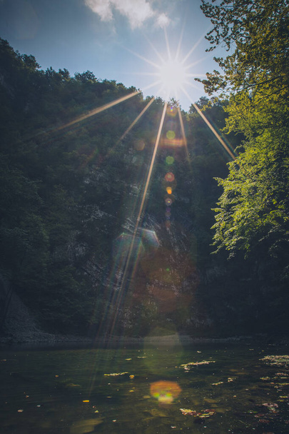 Photo of a small lake called Divje jezero in Slovenia with strong sun flare from the sun above the lake and high vertical rock formation. - Foto, Imagem