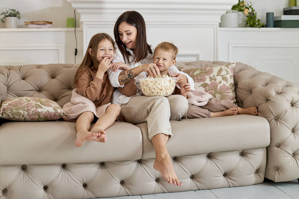 Äiti istuu sohvalla poikansa ja tyttärensä kanssa ja katsoo elokuvaa. Nainen, poika ja tyttö syövät popcornia katsoessaan elokuvaa. Perhe lepää kotona viikonloppuna. - Valokuva, kuva