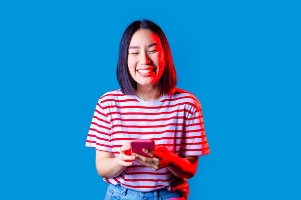 Genç, güzel Asyalı bir kadın mavi arka planda akıllı telefondan mesaj atıyor, gülüyor ve sosyal medyayı ve yeni viral eğilimleri kontrol ederken eğleniyor. - Fotoğraf, Görsel