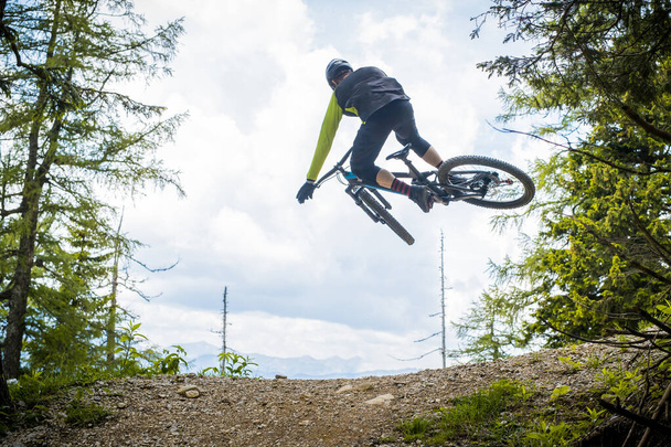 Πίσω πλάνο ενός ποδηλάτη που πηδάει πάνω από ένα χωματόδρομο σε ένα πάρκο ποδηλάτων, που περιβάλλεται από δάσος και δέντρα. Πράσινο ποδηλάτης βουνού σε ένα πράσινο περιβάλλον που εκτελεί ένα μαστίγιο ουρά σε ένα διπλό άλμα. - Φωτογραφία, εικόνα