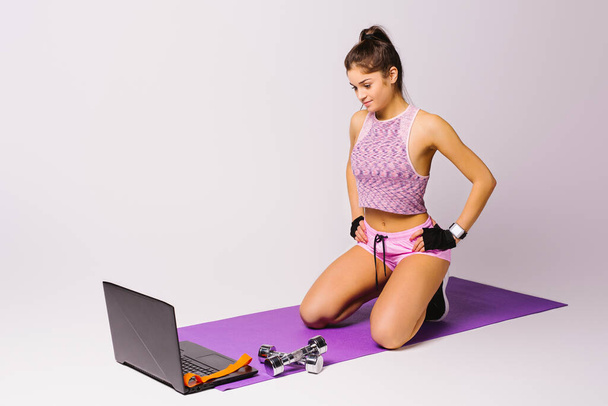 A sportive girl sits on the floor with dumbbells and uses a laptop. Online training concept. White background. Empty space. - Φωτογραφία, εικόνα