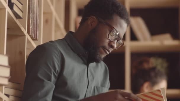 Kippmittelaufnahme eines jungen schwarzen Mannes mit Bart, Brille, der in einem Hipster-Café sitzt, Buch aus dem Regal nimmt, es öffnet und mit Interesse zu lesen beginnt und Kundin im Hintergrund liest - Filmmaterial, Video