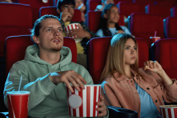 Young couple having popcorn while watching movie together, sitting in cinema auditorium - Valokuva, kuva