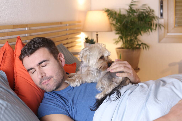 όμορφος νεαρός άνδρας με το σκύλο του να χαλαρώνει στο κρεβάτι στο σπίτι - Φωτογραφία, εικόνα