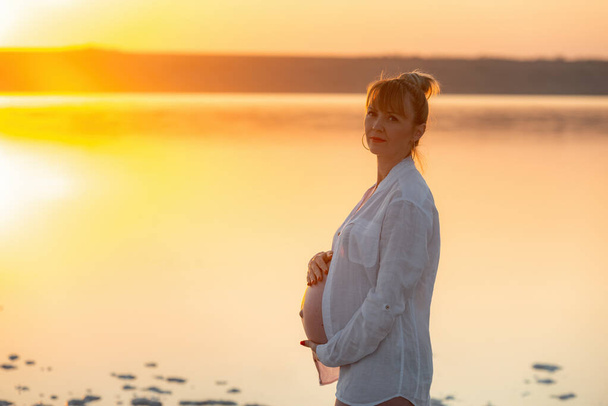 Беременная девушка на закате, беременная женщина отдыхает на пляже. Смотреть в камеру - Фото, изображение