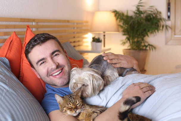 красивый молодой человек с собакой и котом, отдыхающий в постели дома - Фото, изображение