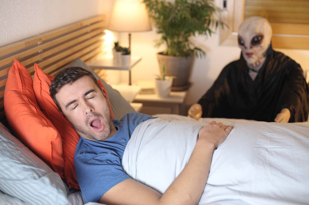 jóképű fiatalember fekszik az ágyban otthon, míg mániákus horror maszk ül a háttérben - Fotó, kép