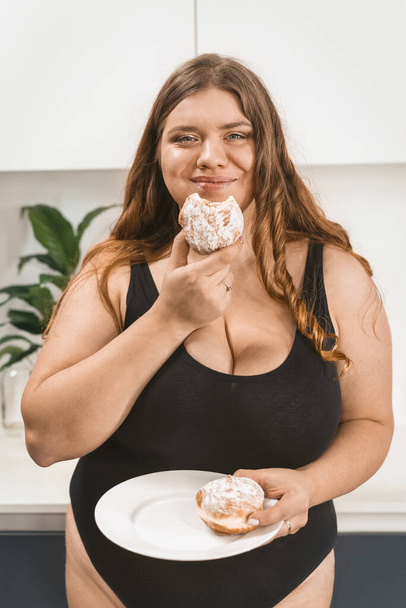Satisfied young fat woman eating a cake happy smiling on camera wearing black swimsuit. Beautiful chubby young woman eating unhealthy food. Fat girl eating cake standing on modern kitchen - Foto, Bild