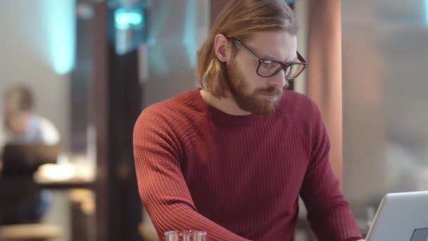 Kippende Taillenaufnahme eines fokussierten jungen kaukasischen Mannes in lässigem Pullover und Brille, der am Tisch im Café sitzt, auf dem Laptop tippt, konzentriert auf den Bildschirm blickt und dann zum Fenster wegschaut. - Filmmaterial, Video