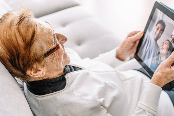 Magas látószögű oldalnézetben elégedett idős nő pihen a kanapén otthon, és miután videohívás a családdal használata közben tabletta - Fotó, kép