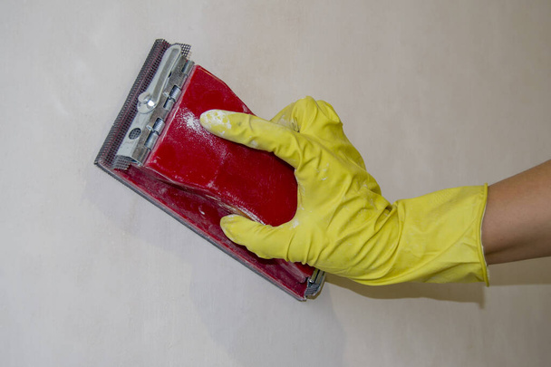 Duvarı zımparalıyorum. Yolsuzlukların ortadan kaldırılması. Bir işçinin elindeki sarı eldiven. kırmızı enstruman - Fotoğraf, Görsel