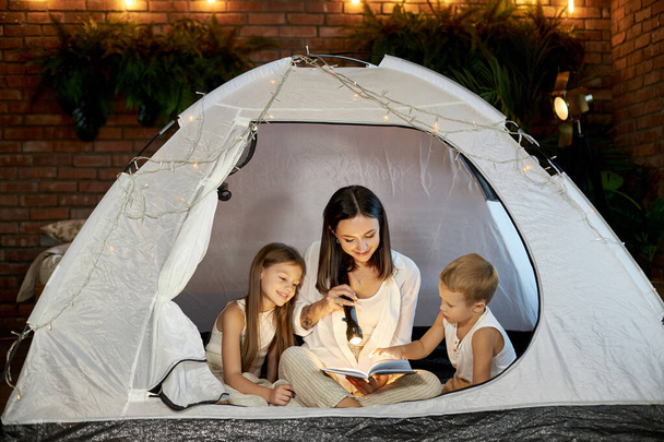 Mom reads children a bedtime story sitting in a tent at home. Mother son and daughter hug and read a book with a flashlight in their hands - Fotoğraf, Görsel