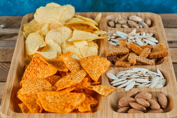 Plate of assorted snacks on a wooden table. Chips, crackers, almonds, pistachios, sunflower seeds - Photo, Image