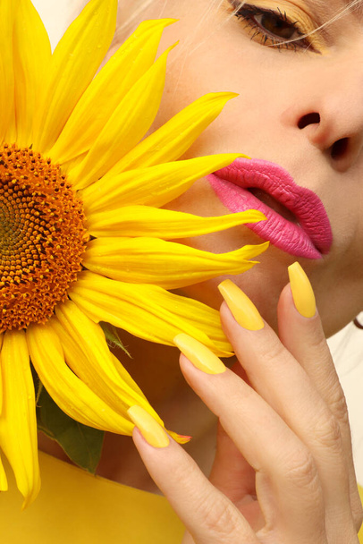 Fashionable manicure on long nails covered with yellow nail polish on a woman with a sunflower. - Photo, Image