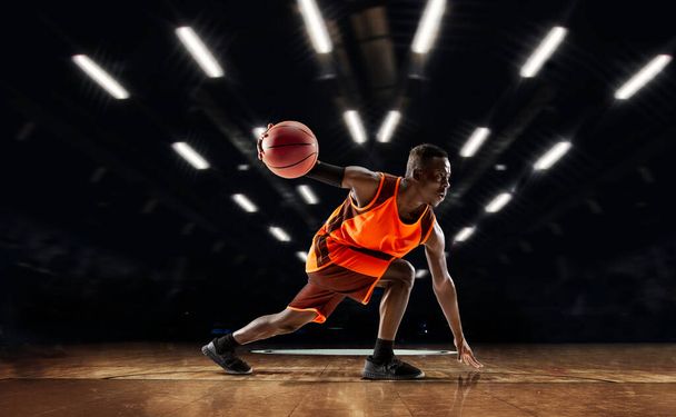 African-american young basketball player in action and flashlights over gym background. Concept of sport, movement, energy and dynamic, healthy lifestyle. - Фото, изображение