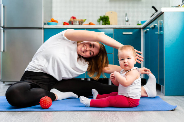 A young happy mother is doing yoga on a mat in the kitchen, and her baby is sitting next to her. The concept of home sports training with children. - Photo, Image