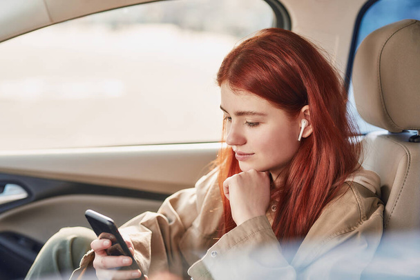 Teenage girl in earphones looking at the screen with a smile, while using her phone, watching videos sitting in the car - Photo, Image