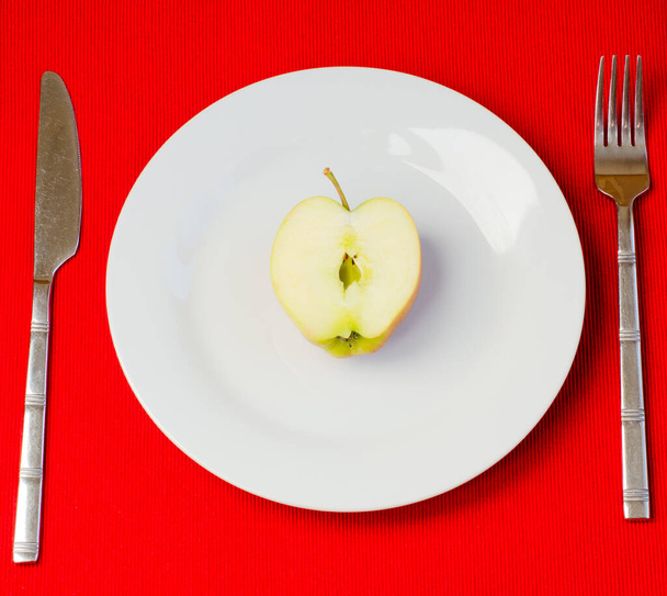 Яблоко разрезано пополам на белой тарелке со столовыми приборами с обеих сторон на красной текстурированной скатерти - Фото, изображение