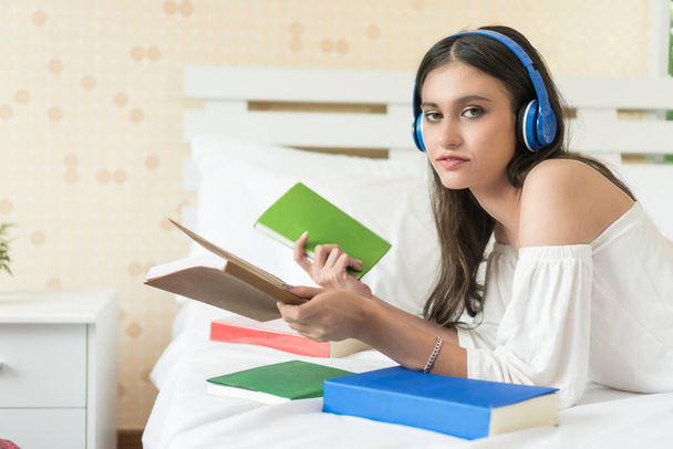 Beautiful smiling young woman lying on bed wearing blue headphones, holding a book in her hand - Foto, Imagem