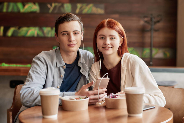 Happy teenage couple smiling at camera while listening to music together using smartphone and same pair of earphones, sitting in a cafe on a daytime - Valokuva, kuva