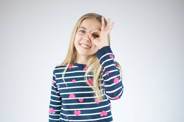 Little girl in a striped shirt on a white background. The child shows an okay sign. The concept of success, approval. - Foto, imagen