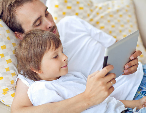 Γιος και ο πατέρας παίζουν ένα παιχνίδι υπολογιστή σε ένα tablet Closeup του νεαρού αγοριού κάθεται με τον πατέρα και χρησιμοποιώντας το κινητό τηλέφωνο στο σύγχρονο ηλιόλουστο μέρος. - Φωτογραφία, εικόνα