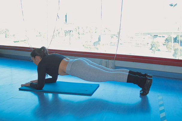 Brunette woman doing abdominal plank in gym, in front of a large window and with blue parquet floor. - Photo, Image