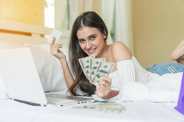 Schöne lächelnde junge Frau liegt im Bett und hält Dollar und Kreditkarte mit einem Laptop vor sich, um online einzukaufen. - Foto, Bild