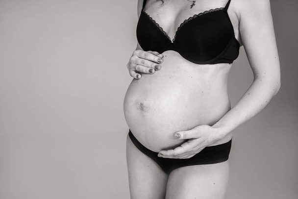 Una chica embarazada en lencería negra sostiene una panza. Mujer esperando el nacimiento de un niño - Foto, Imagen